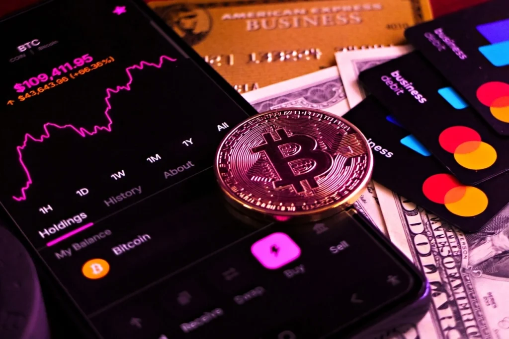 Bitcoin trading interface on smartphone with physical Bitcoin coin and credit cards in vibrant pink and purple lighting, illustrating Bitcoin prices.