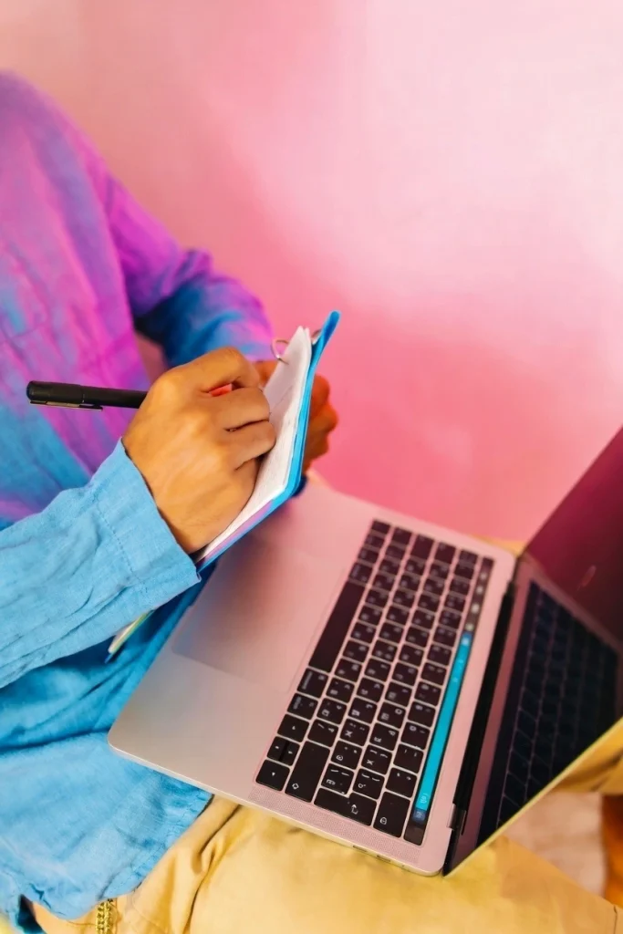 How Simulated Crypto Trading Works: Person taking notes in a notebook while studying on a laptop, representing the discipline and learning process required for beginners in simulated crypto trading.